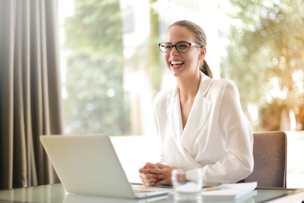 Frau sitzt vor geöffnetem Laptop, lacht und trägt ein elegantes Büro-Outfit